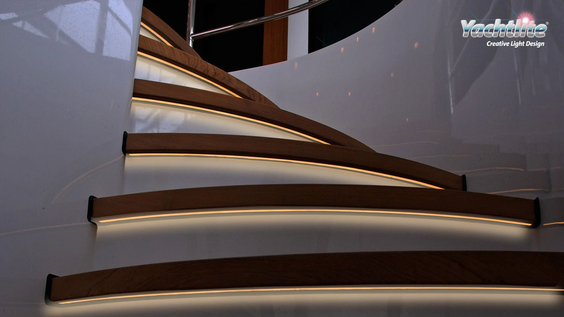 Curved wooden yacht staircase illuminated with warm white Yachtlite FormLite LED lighting integrated beneath each step. The soft ambient glow highlights the modern staircase design, polished white walls, and luxury interior details, creating a sleek and elegant onboard atmosphere. Yachtlite “Creative Light Design” logo appears in the upper right corner.