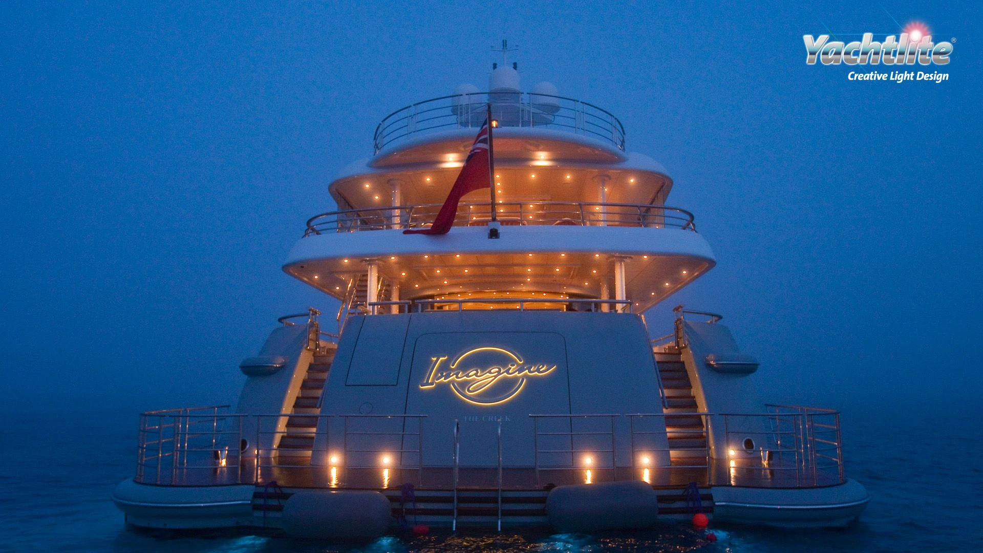 The back of a superyacht at dusk, showing its yacht sign which has gold lighting.