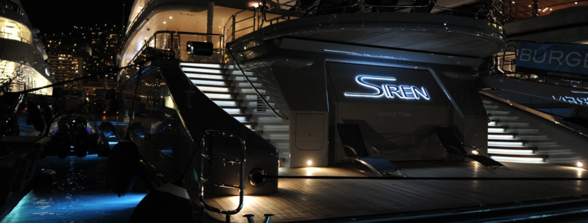 The stern of Siren, a yacht at night in a marina, featuring ambient step lighting and an illuminated yacht sign.