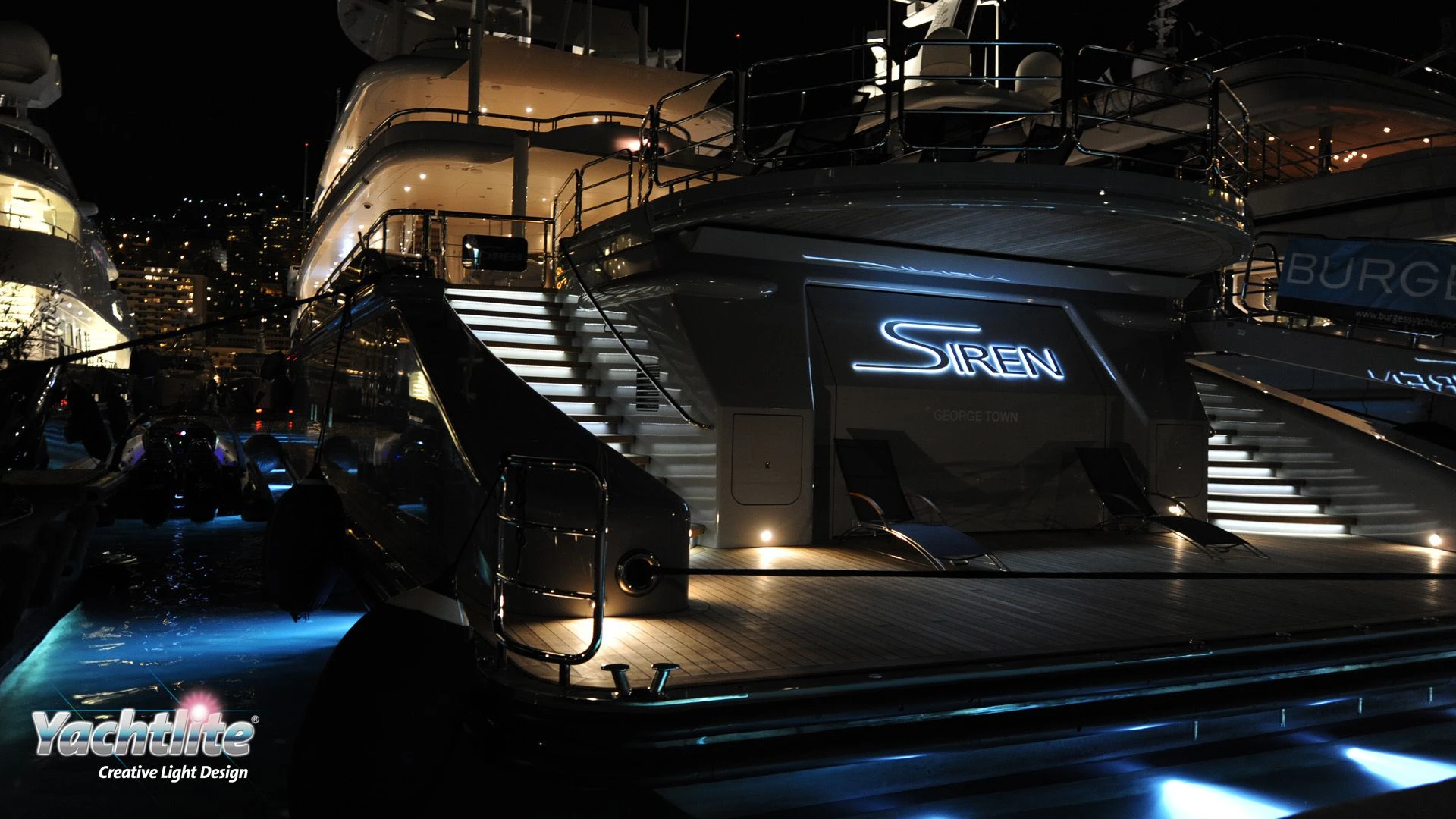The stern of Siren, a yacht at night in a marina, featuring ambient step lighting and an illuminated yacht sign.