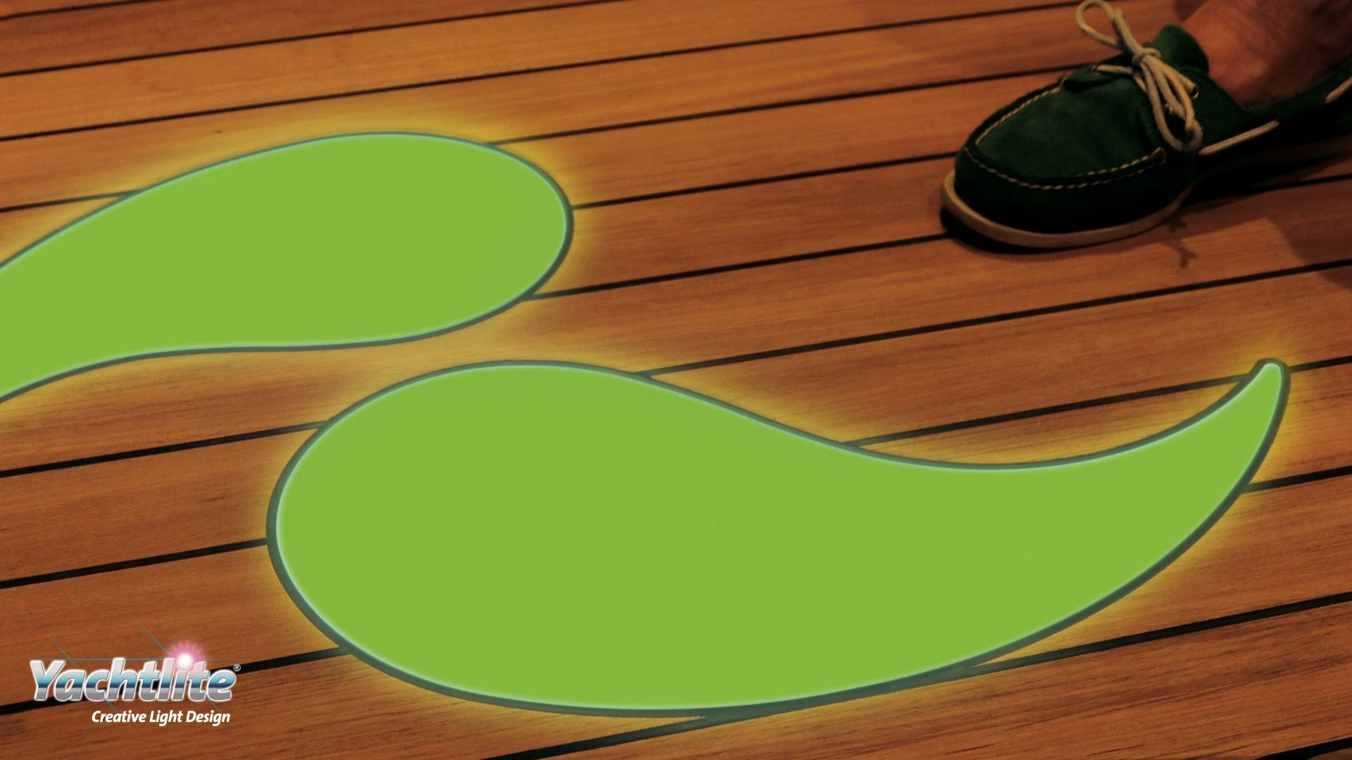 Two illuminated green curved shapes are embedded into a wooden yacht deck. A person's foot is visible in the top-right corner.